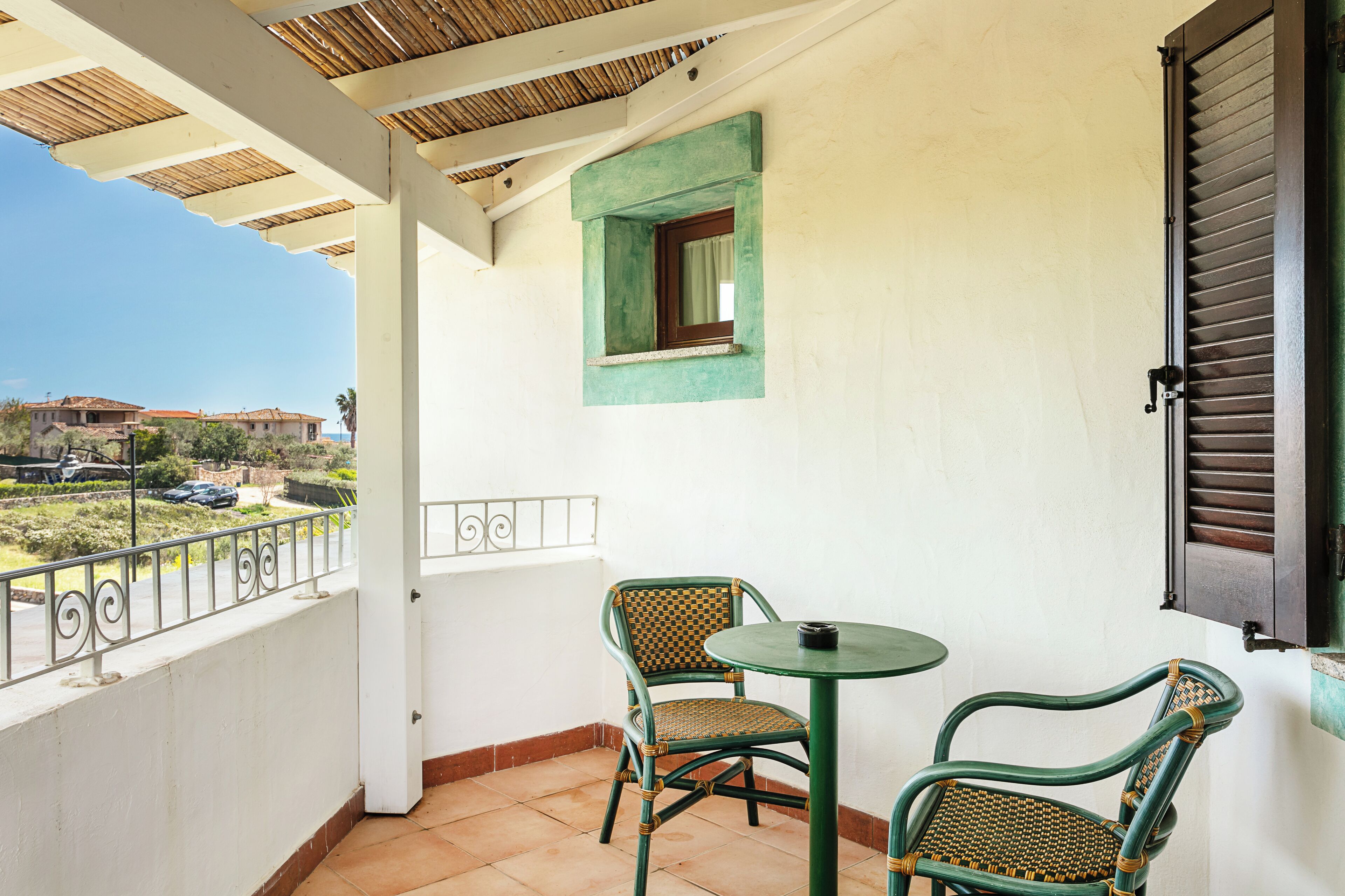 A balcony with two wicker chairs and a small table, a window with a green frame, and a view of buildings and a car in the background.