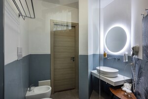Un baño con puerta de madera, un lavabo blanco, un espejo con luz circular y un perchero para toallas.