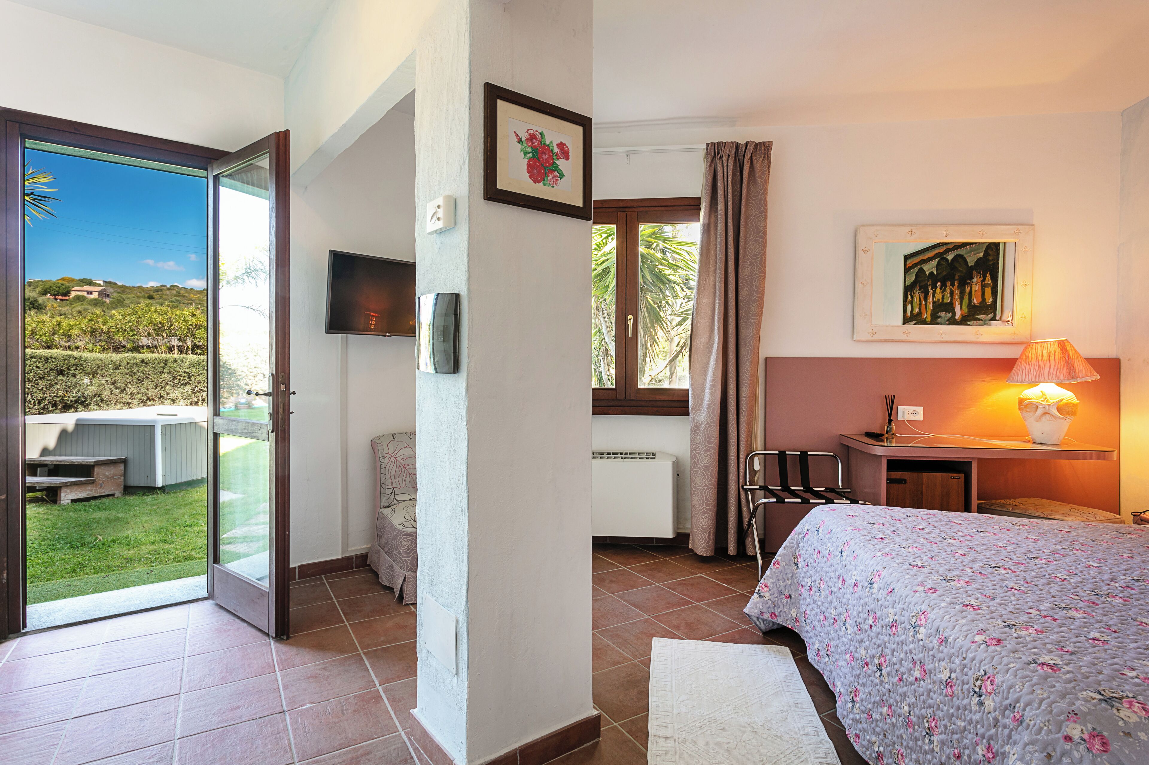 Ein Schlafzimmer mit Bett, Fernseher, Fenster mit Vorhängen, Heizkörper und Blick ins Freie.