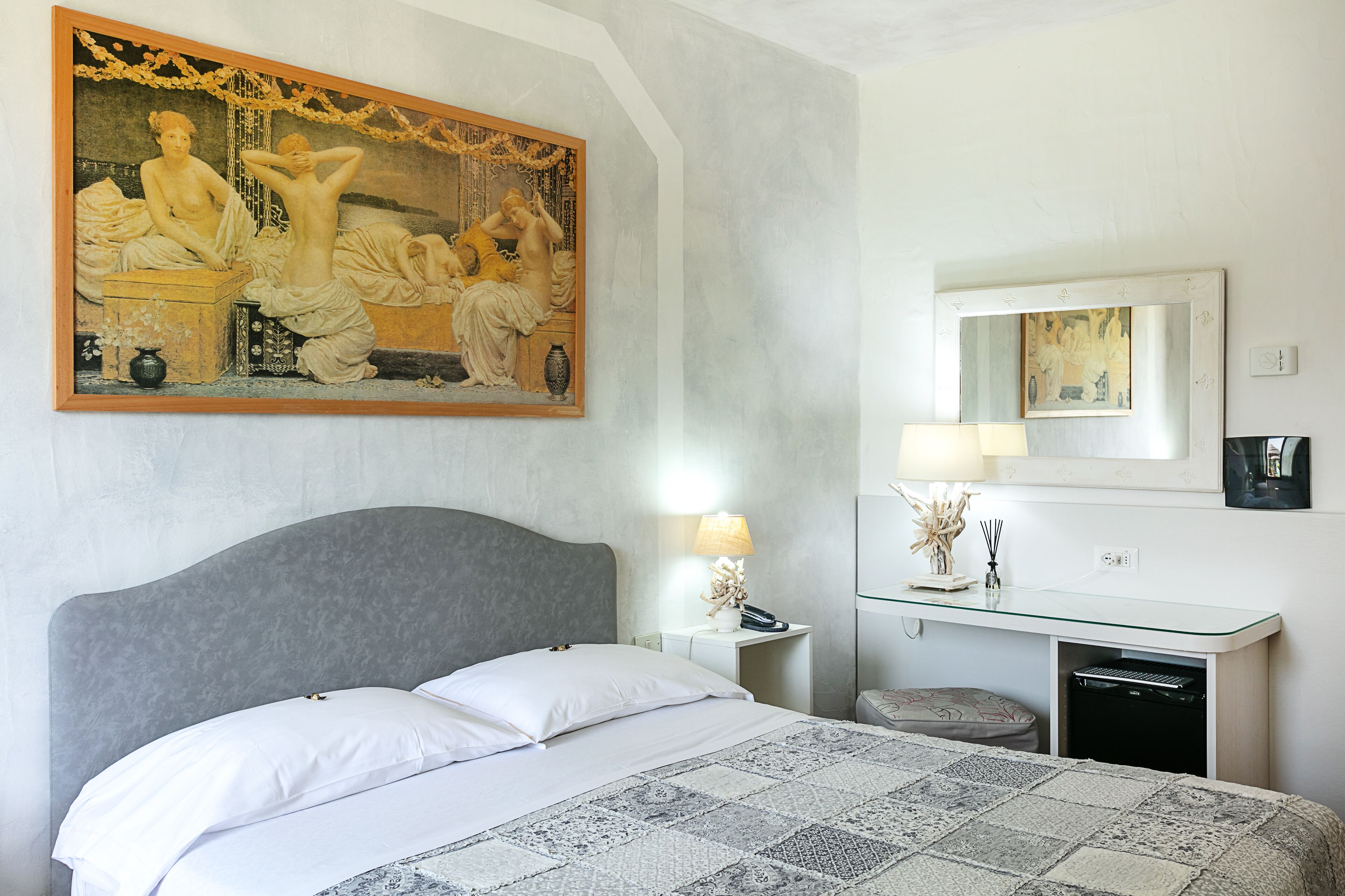 Habitación de hotel con un cuadro clásico grande, una cama con sábanas blancas, una mesita de noche con lámpara y un escritorio pequeño con espejo y lámpara.