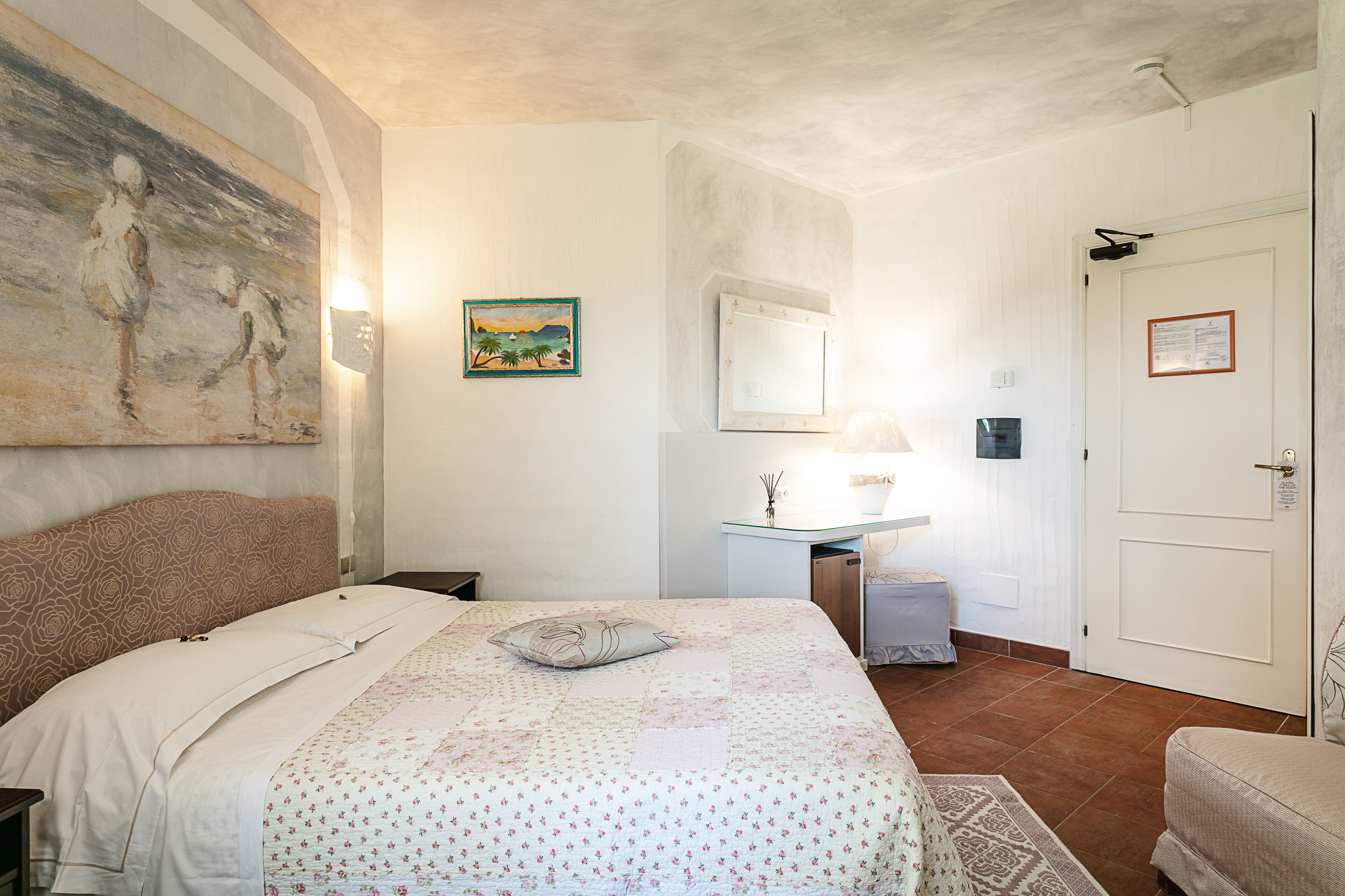 Un dormitorio con cama, mesita de noche, escritorio, silla, puerta y un cuadro en la pared.