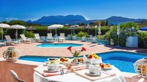 Un área de comedor junto a la piscina con una mesa preparada para el desayuno, con vistas a una piscina y un paisaje que incluye montañas y el mar.