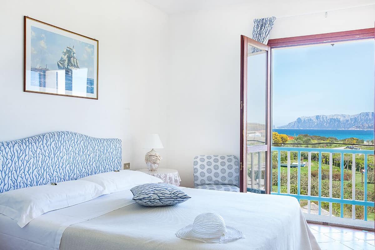 Un dormitorio con cama, mesita de noche, lámpara, silla y un balcón con vistas al mar y a las montañas.