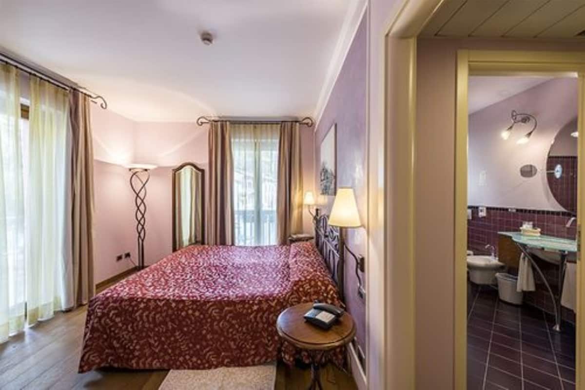 Habitación de hotel con una cama grande, una mesita de noche con lámpara, un espejo y vista al baño.