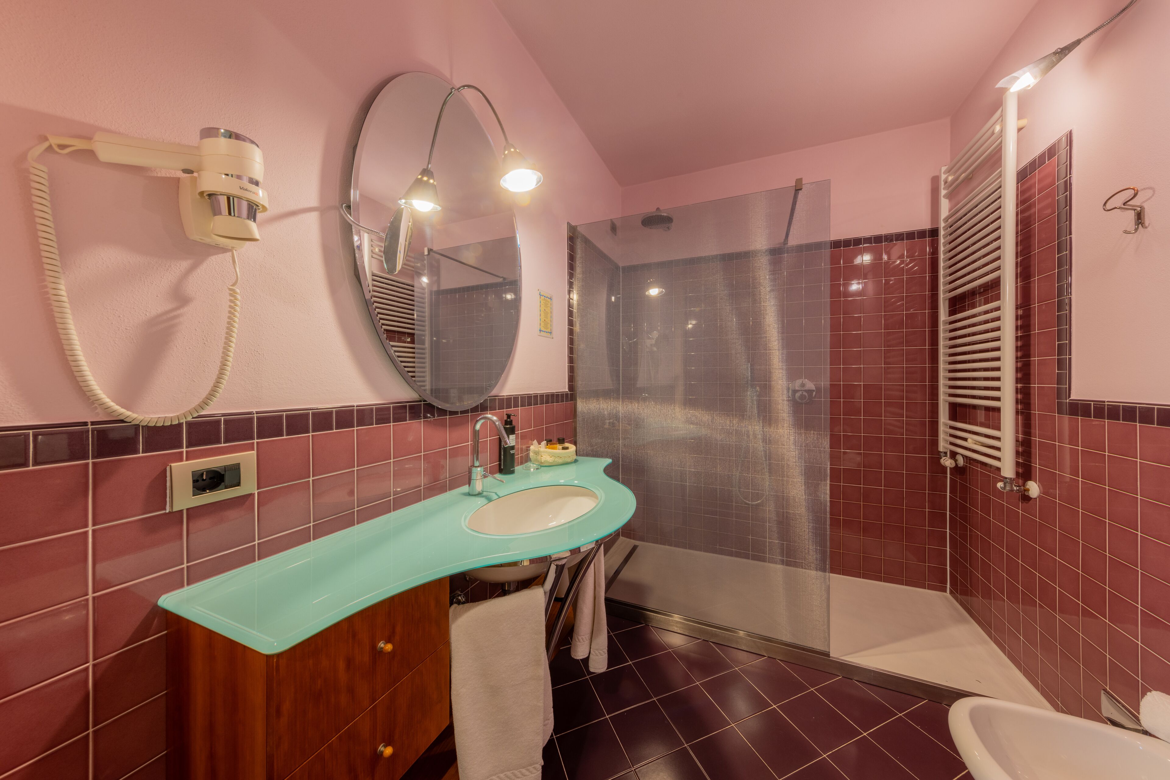 Un baño con pared de azulejos rosa y rojos, un lavamanos con encimera verde, un espejo, un secador de pelo y una zona de ducha.