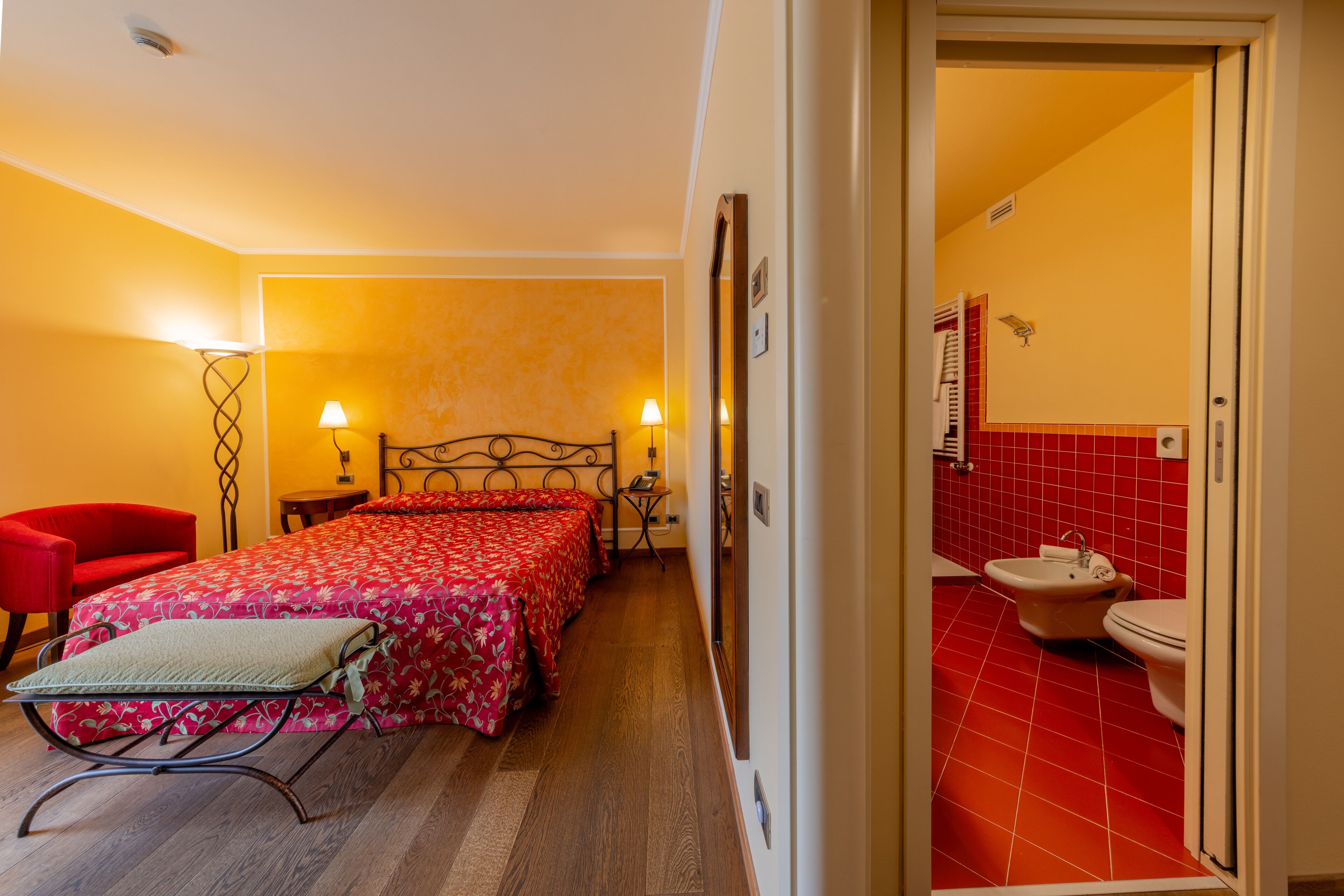 A hotel room with a bed, bedside tables, a red chair, and a bathroom with red tiles.
