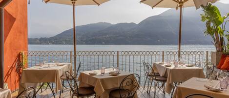 An outdoor dining area with tables and chairs, umbrellas, and a view of a lake and mountains.