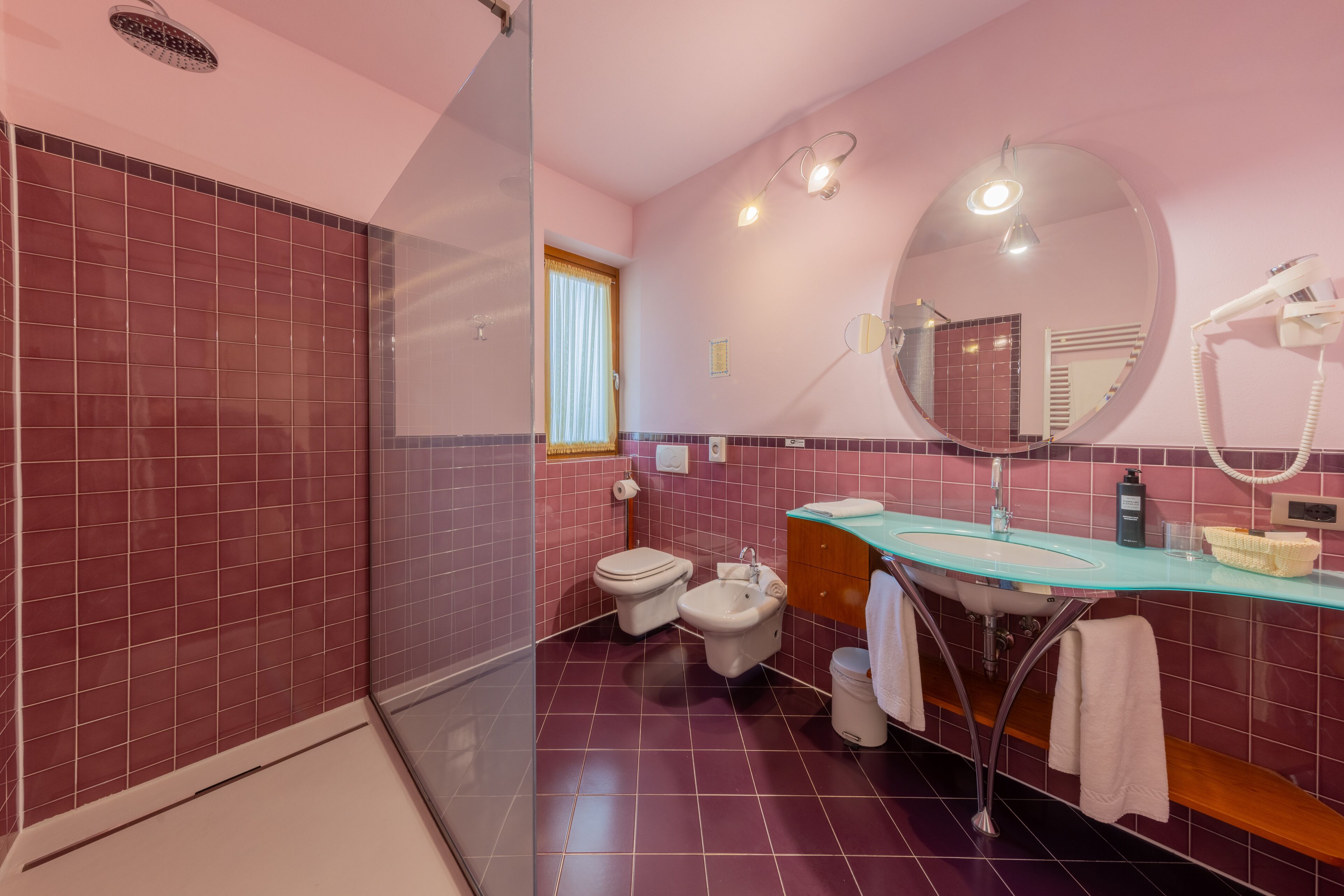 A bathroom with a glass shower enclosure, a toilet, a bidet, and a vanity with a mirror and a hairdryer.