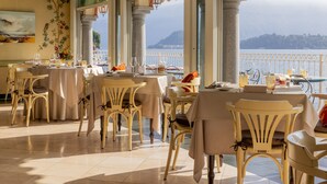 A dining area with tables and chairs, large windows with a view of a body of water and hills, and a decorative wall with a painting. - La Darsena Boutique Hotel & Restaurant (Tremezzina)