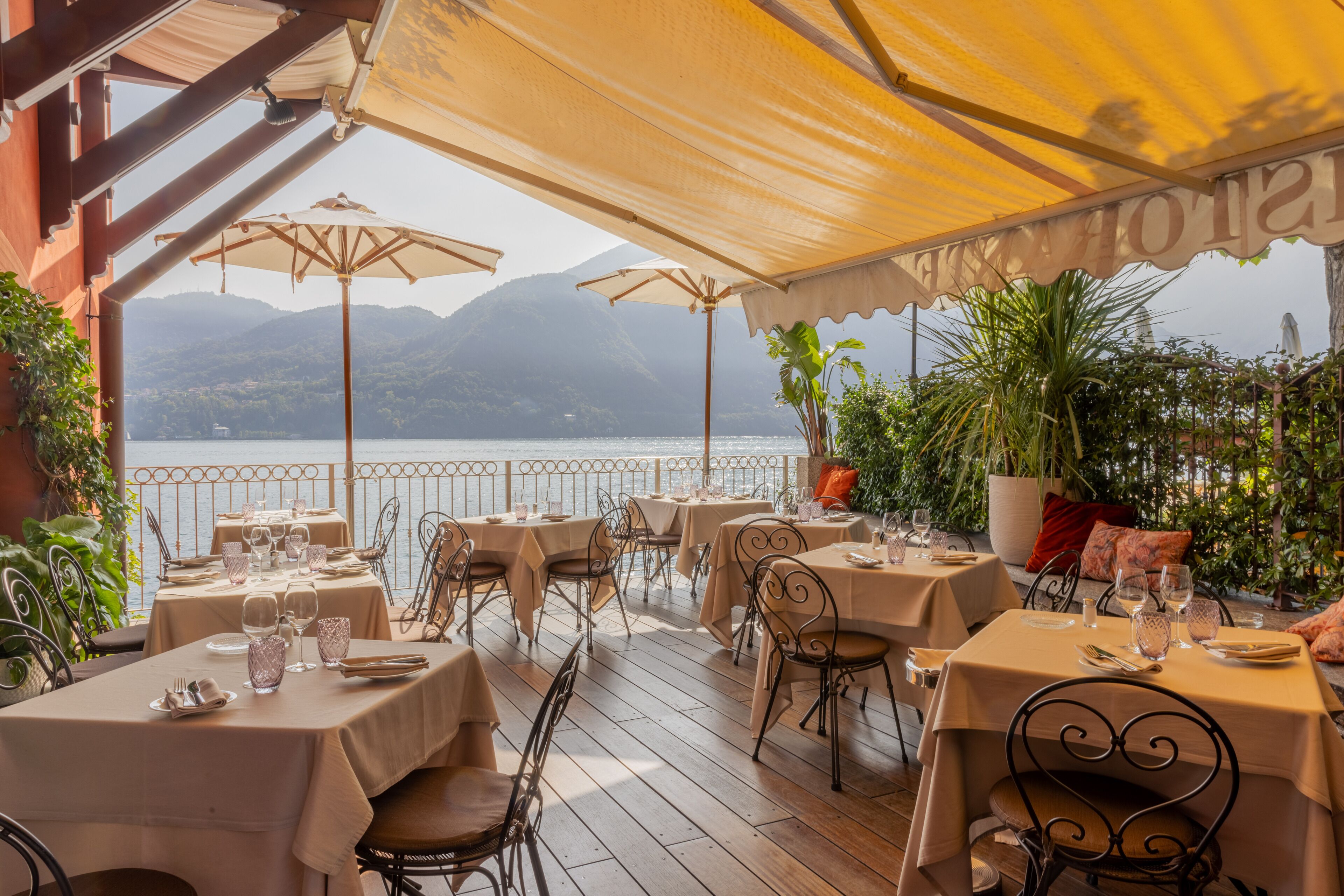 An outdoor dining area with tables set for two, overlooking a body of water and mountains.