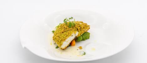 Un plato blanco con un trozo de pescado cubierto por una sustancia amarilla desmenuzable, acompañado de un vegetal verde y una cucharada de salsa.