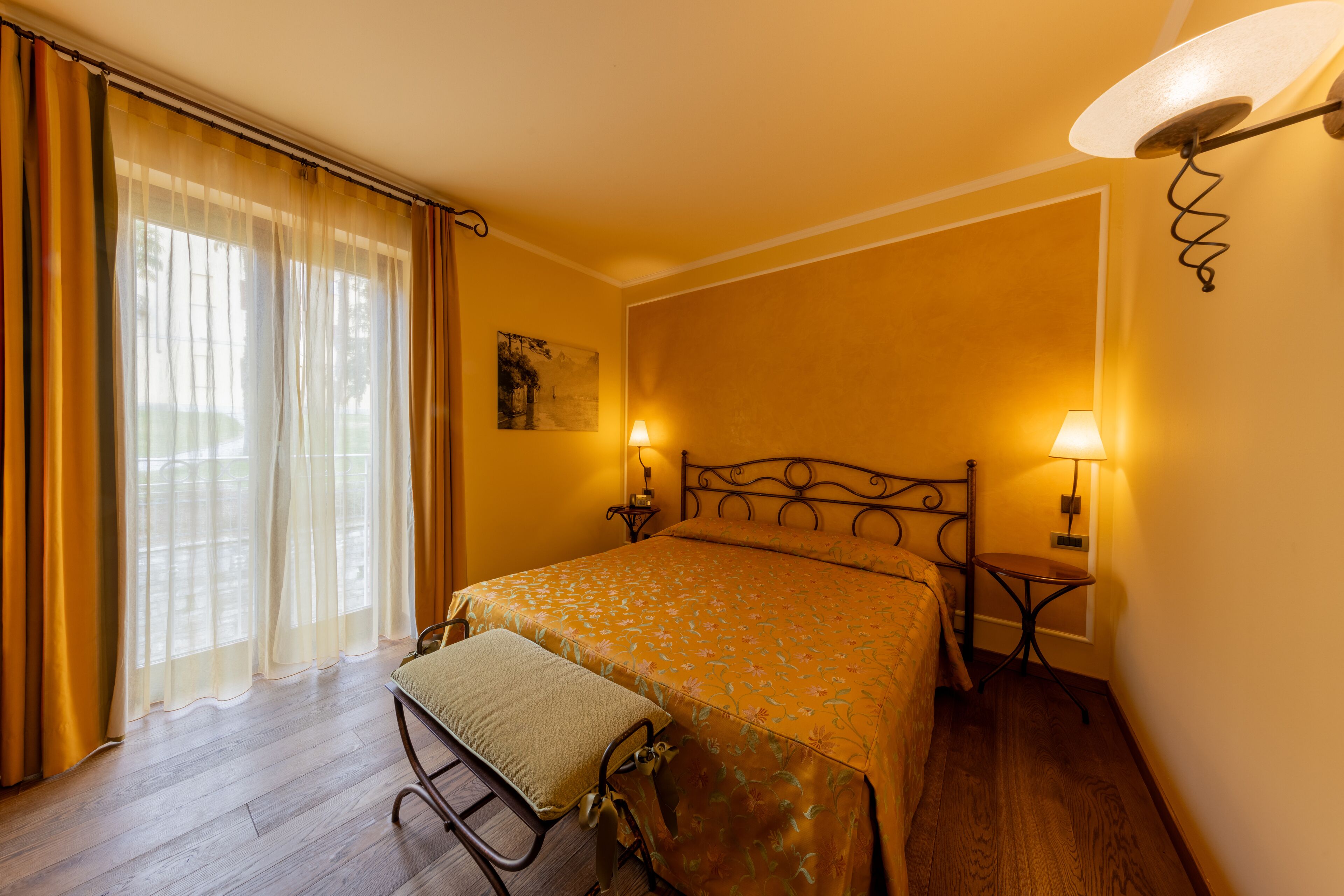 A bedroom with a bed, a bedside table, a lamp, a window with curtains, and a wall-mounted light fixture.