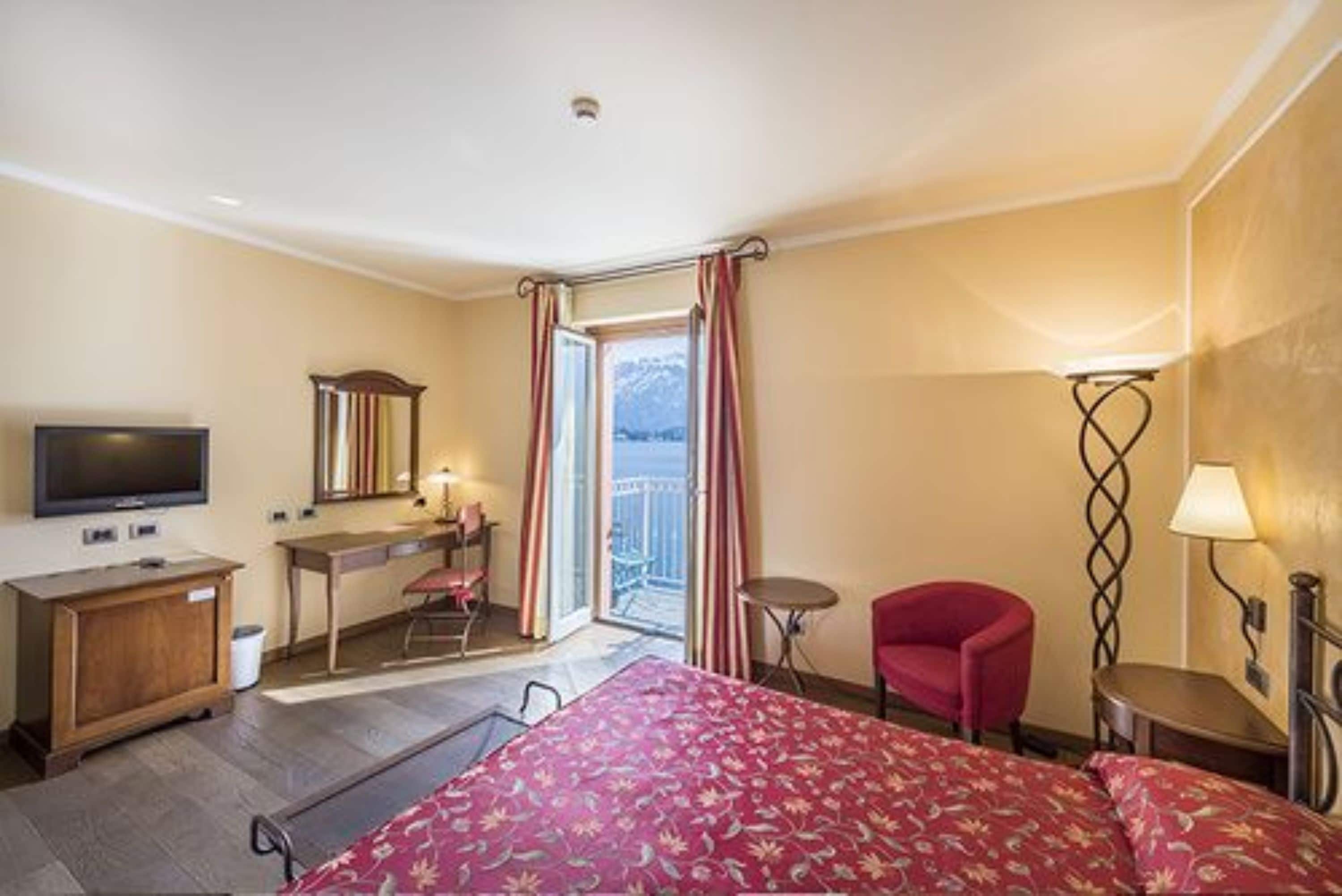 A hotel room with a bed, a desk, a chair, a TV, a balcony with curtains, and a lamp.