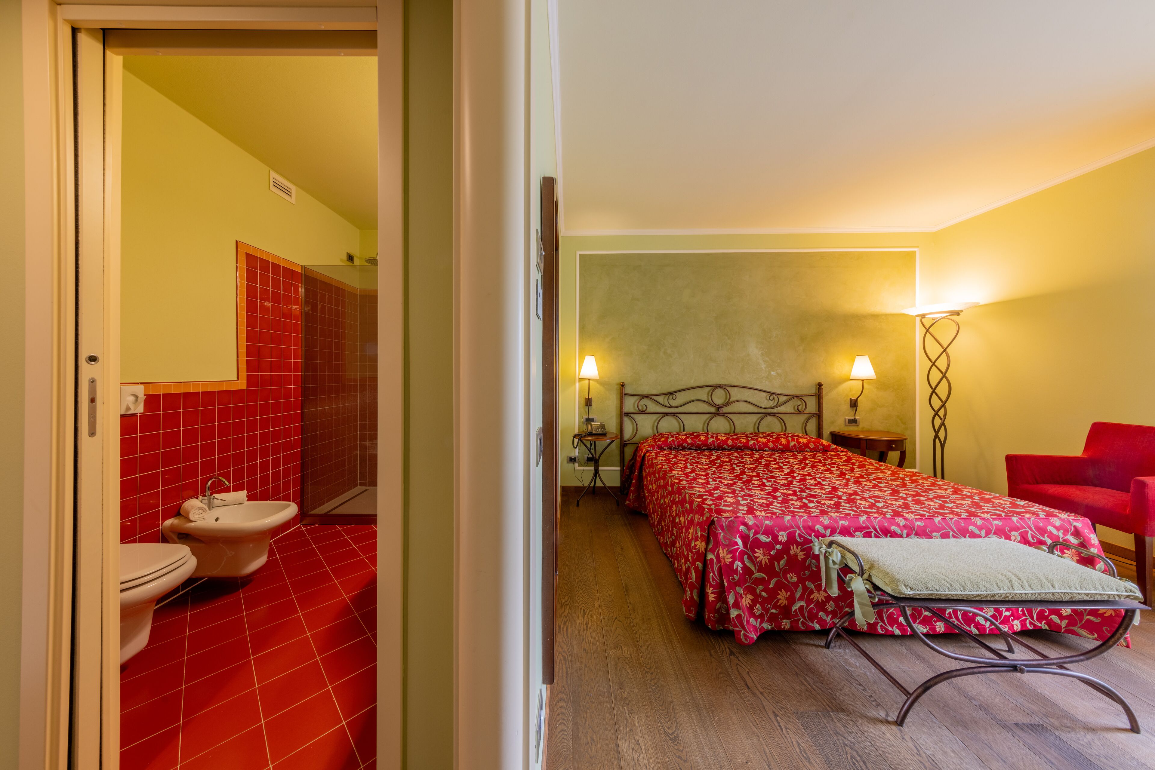 Une chambre d’hôtel avec une salle de bain aux carreaux rouges, un lit recouvert d’une courtepointe à motifs floraux rouges, deux lampes de chevet et une chaise rouge.