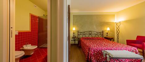 Habitación de hotel con baño de baldosas rojas, cama con colcha floral roja, dos lámparas de noche y una silla roja.