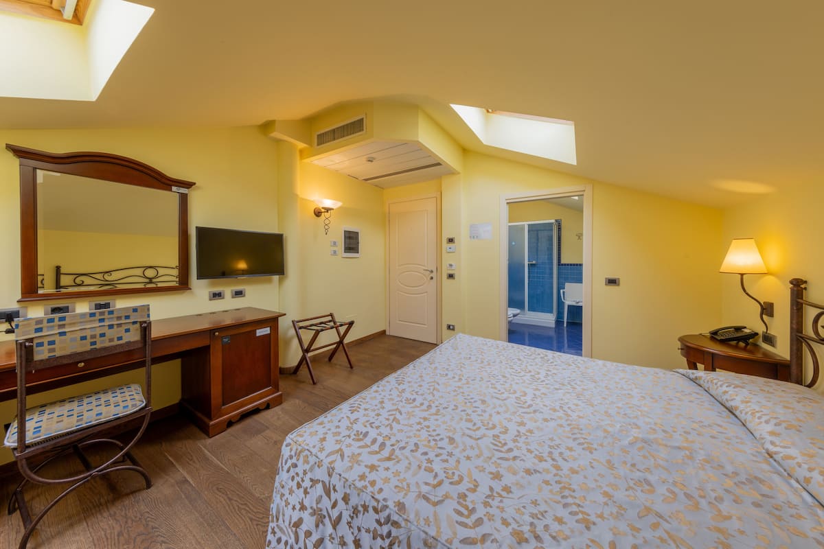 Habitación de hotel con cama, escritorio, silla, espejo y baño visible a través de una puerta abierta.