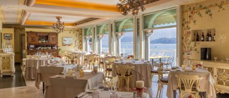 A restaurant with multiple tables, elegant chandeliers, and a view of the sea.