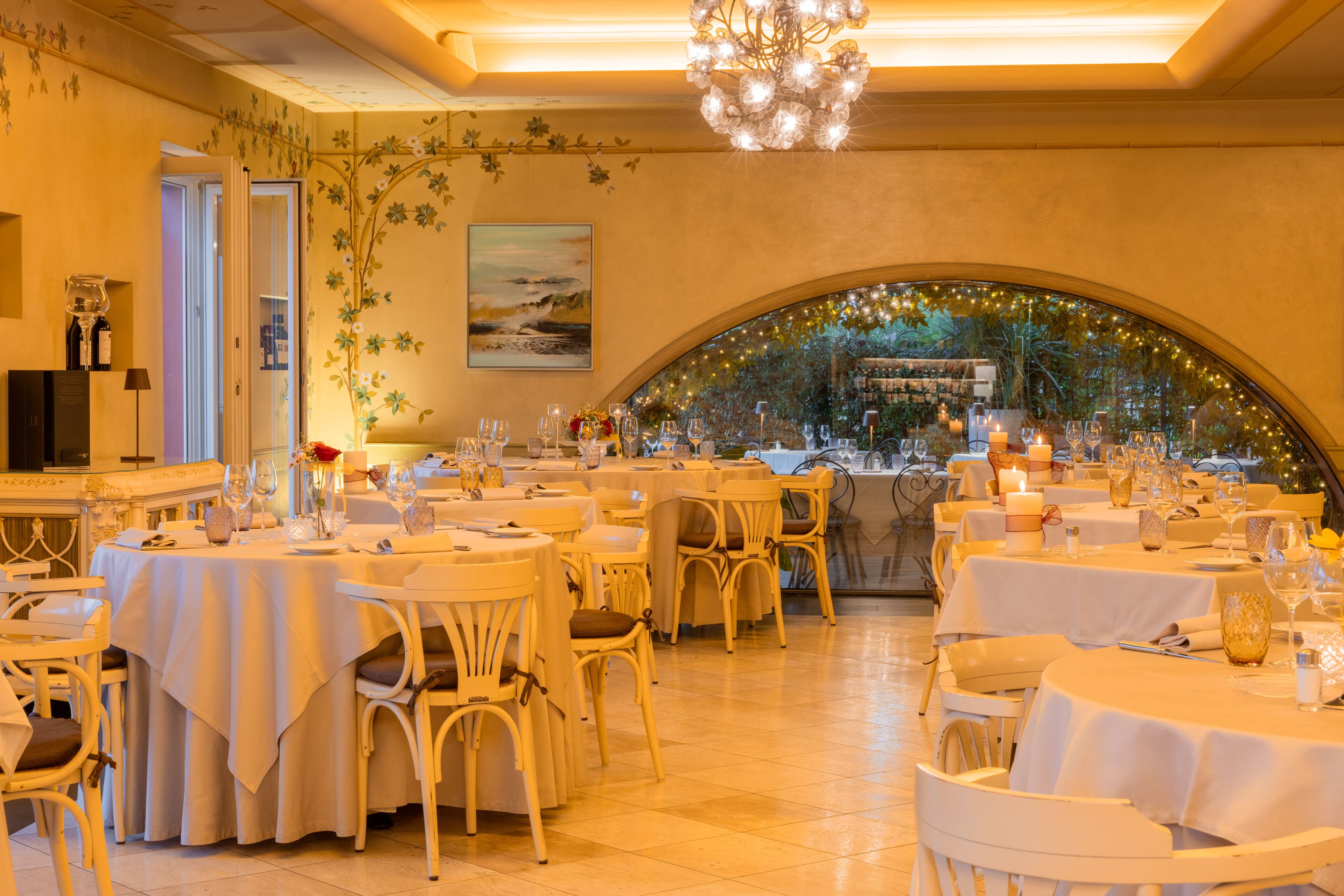 A restaurant with multiple tables set for dining, white chairs, and a chandelier.