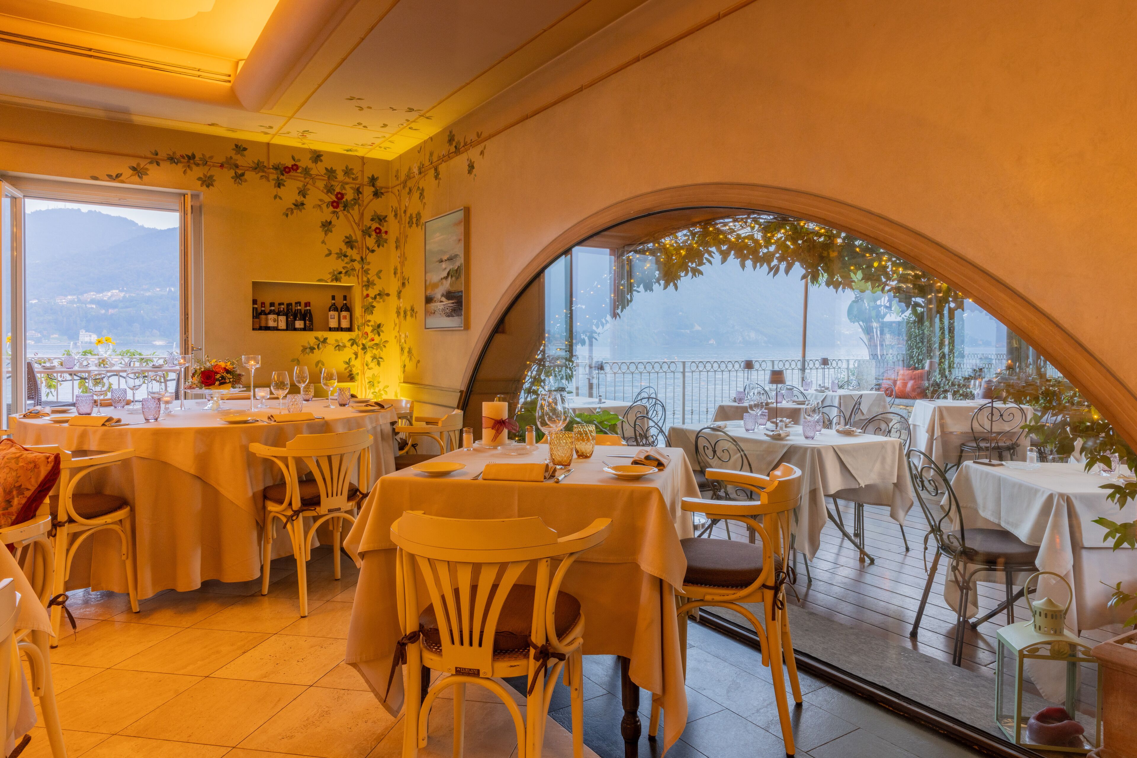 Un restaurante con terraza, mesas y sillas blancas, y vistas a montañas a través de amplios ventanales.