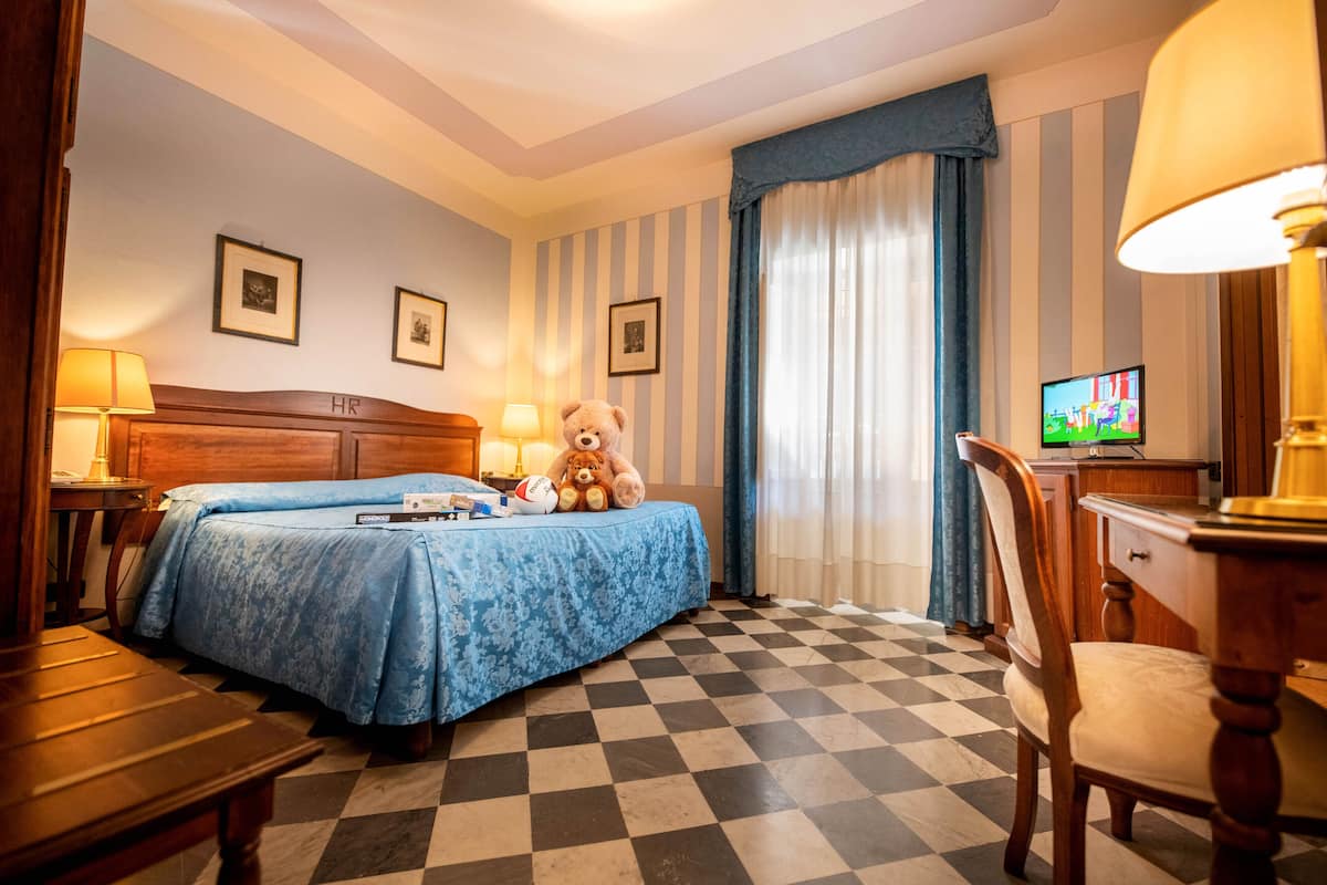 A hotel room with a checkered floor, a bed with a blue bedspread, a teddy bear, a television, and a desk with a lamp.