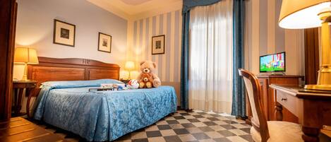 A hotel room with a checkered floor, a bed with a blue bedspread, a teddy bear, a television, and a desk with a lamp.