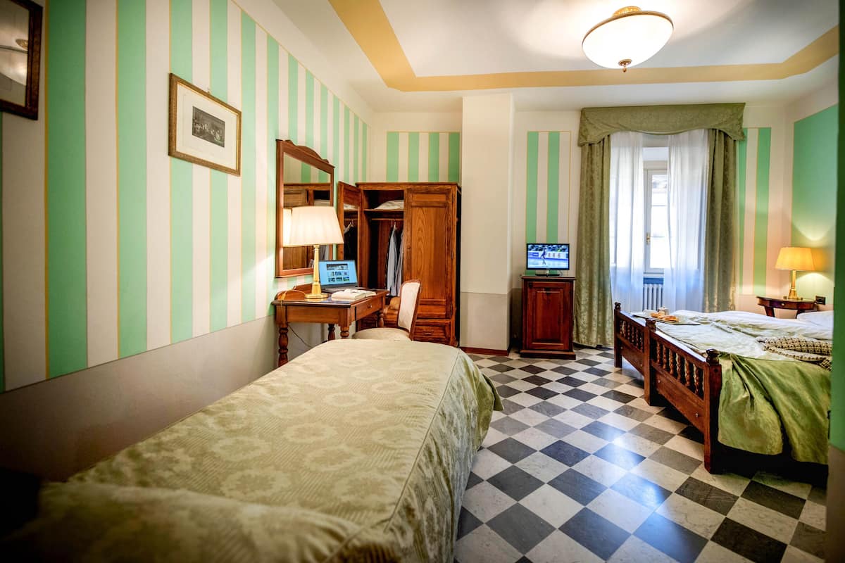 A hotel room with a checkered floor, striped wallpaper, a bed, a desk with a computer, a wardrobe, and a television.