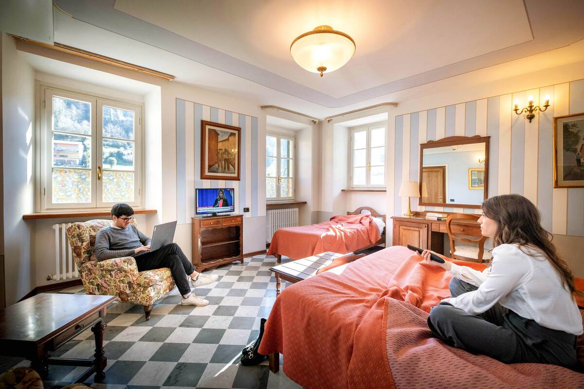 A hotel room with a checkered floor, two beds, a desk with a computer, a chair, and a window with a view.