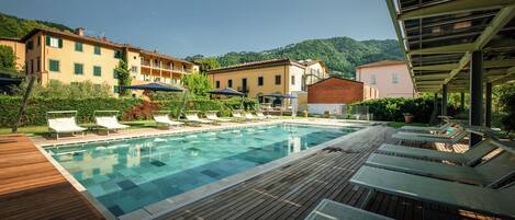 A swimming pool with lounge chairs and umbrellas, surrounded by buildings and greenery.
