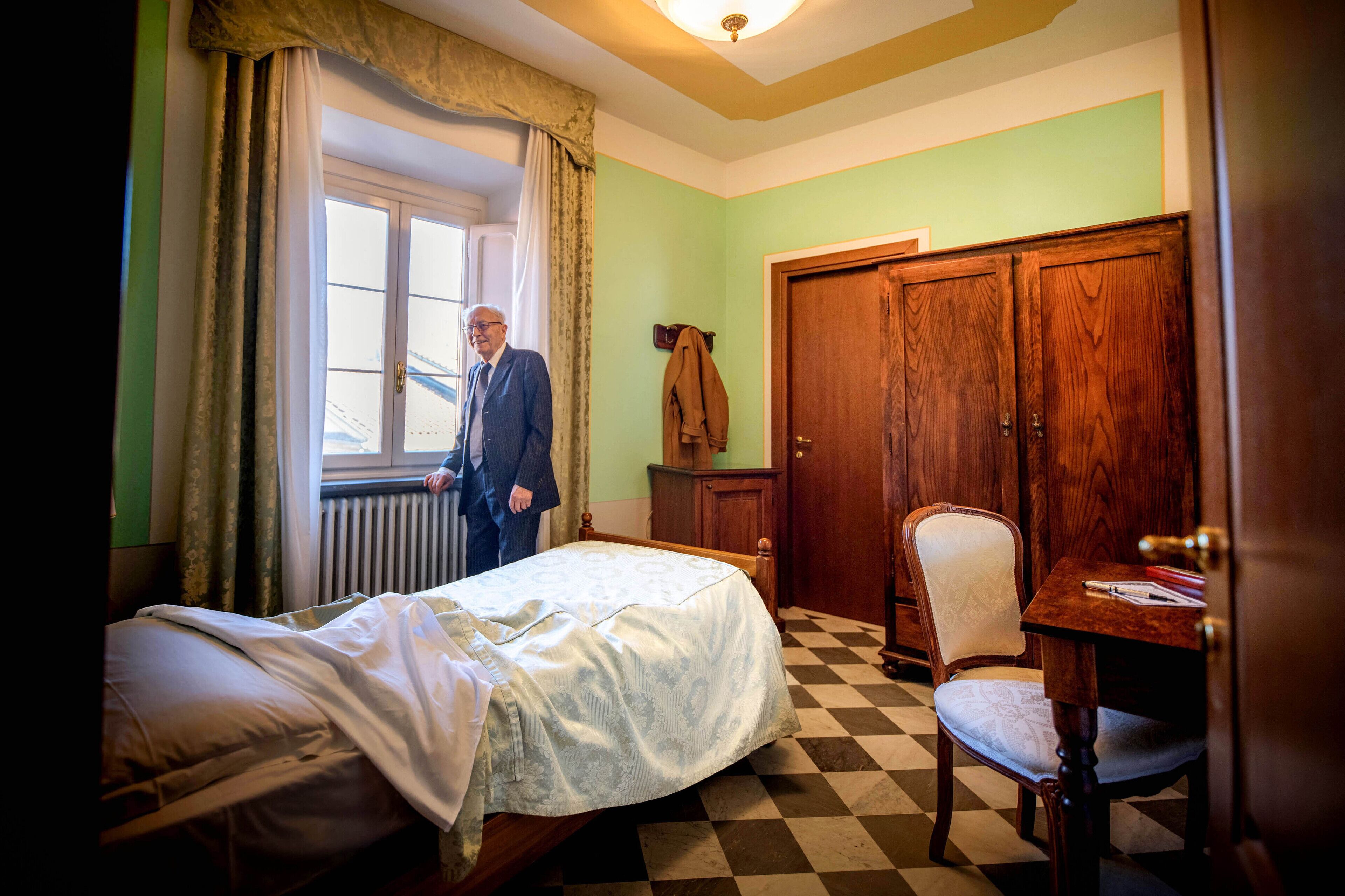 A room with a checkered floor, a bed with a white bedspread, a wooden chair, a wooden wardrobe, and a window with curtains.