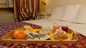 Een hotelkamer met een net opgemaakt bed, een nachtkastje met een lamp, en een dienblad met ontbijtartikelen zoals brood, fruit en drankjes.