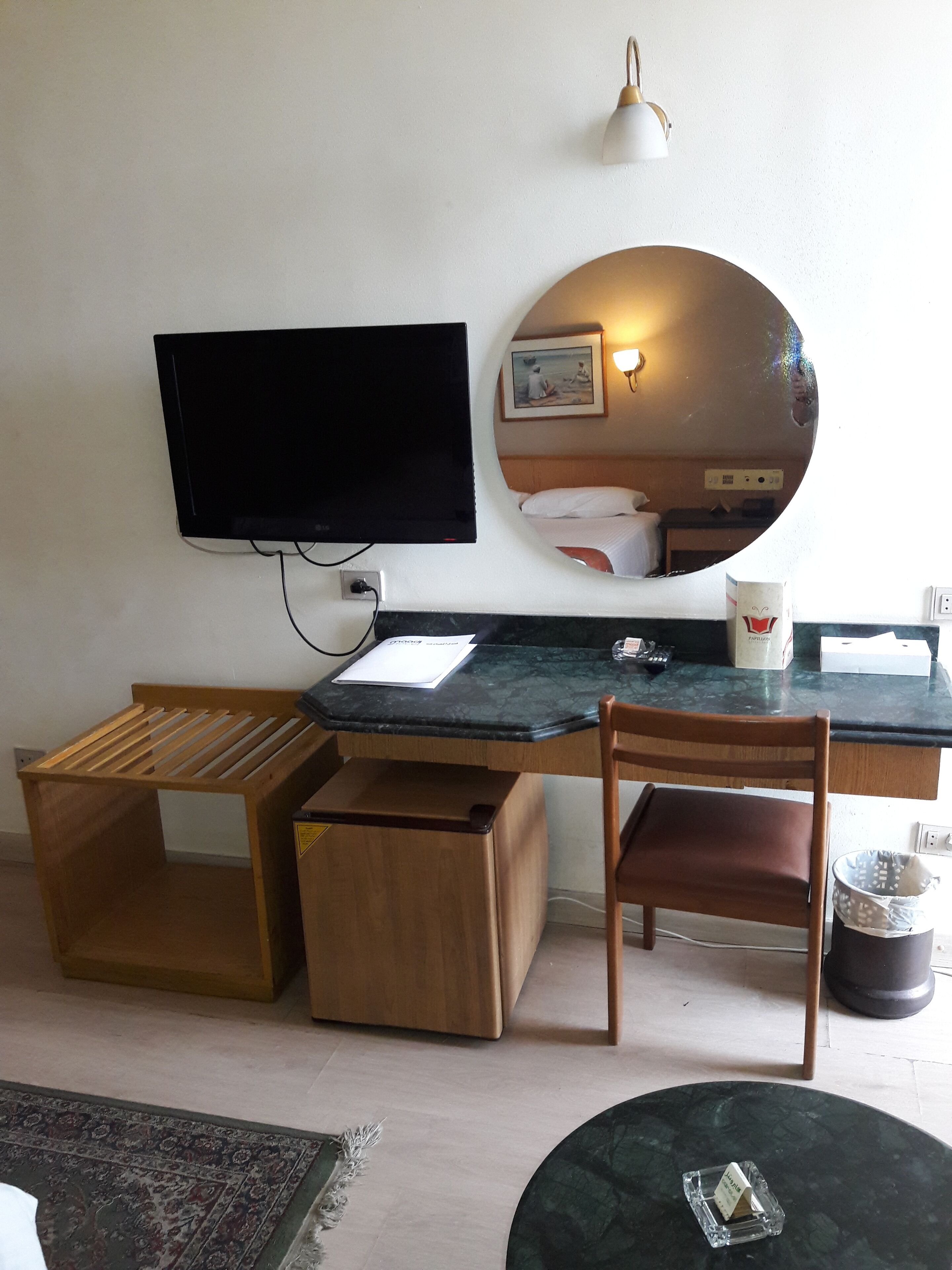A compact hotel room with a wooden desk, a television, a mirror, and a small round table.