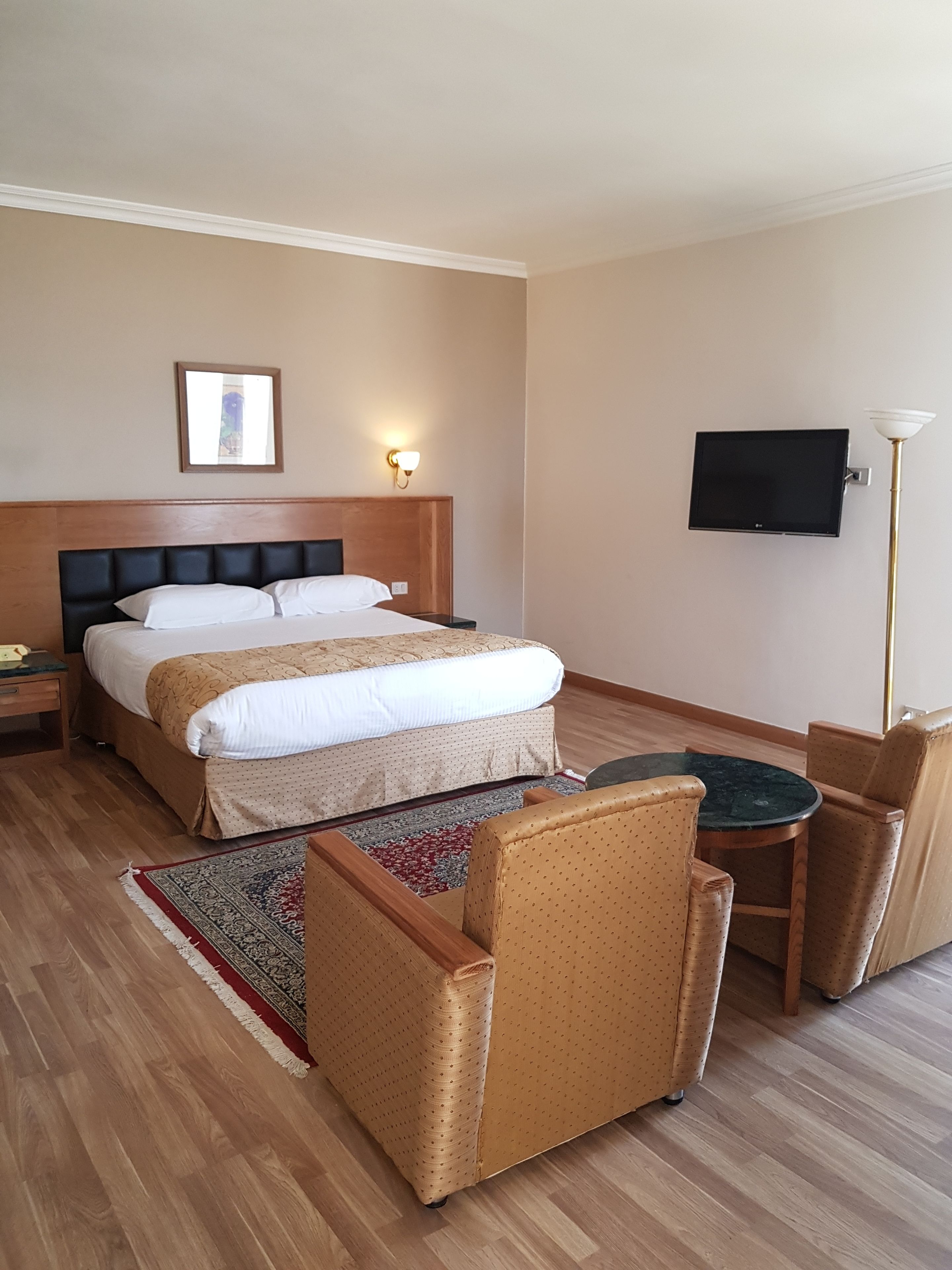 A hotel room with a double bed, a small table with two chairs, a TV on the wall, and a floor lamp.