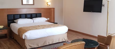 A hotel room with a double bed, a small table with two chairs, a TV on the wall, and a floor lamp.