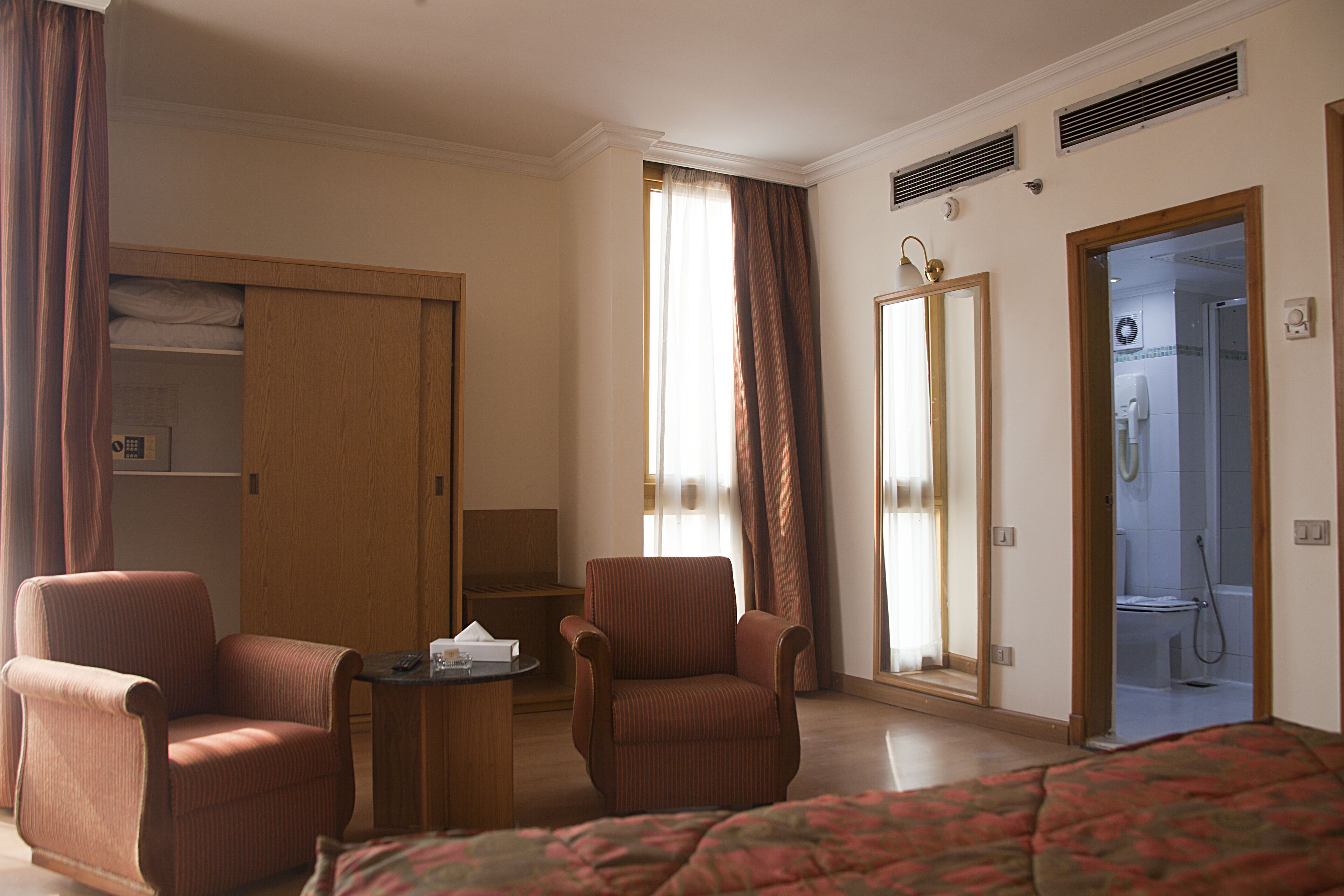 Habitación de hotel con cama, dos sillones, una mesita, armario, espejo y baño visible a través de una puerta abierta.