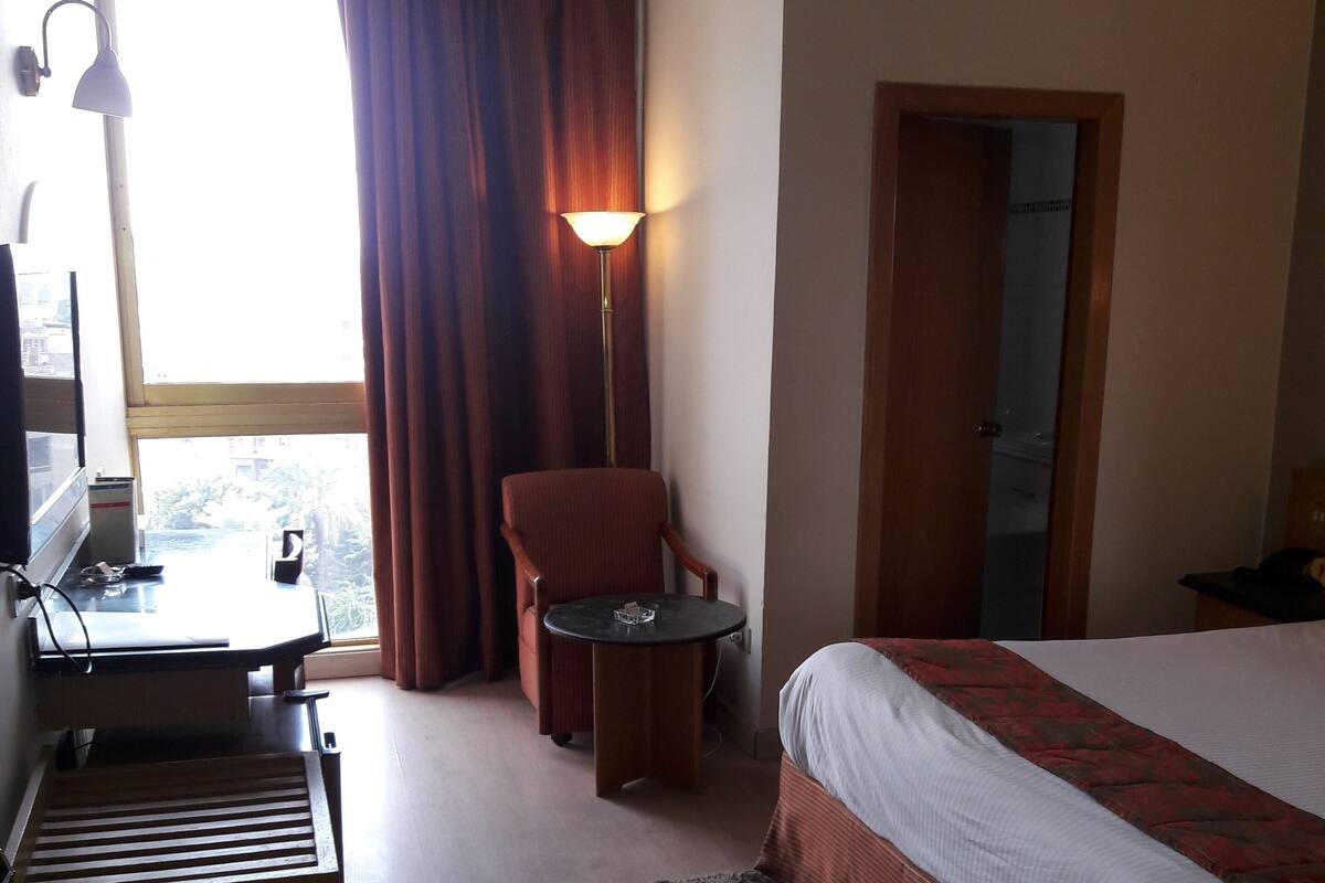 Quarto de hotel com cama, escrivaninha, cadeira, luminária de chão e vista para o exterior.