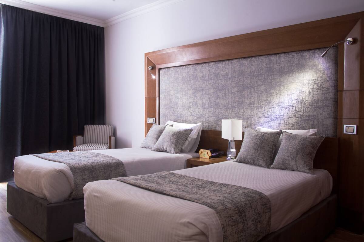Quarto de hotel com duas camas, uma mesa de cabeceira, uma luminária e uma cadeira.