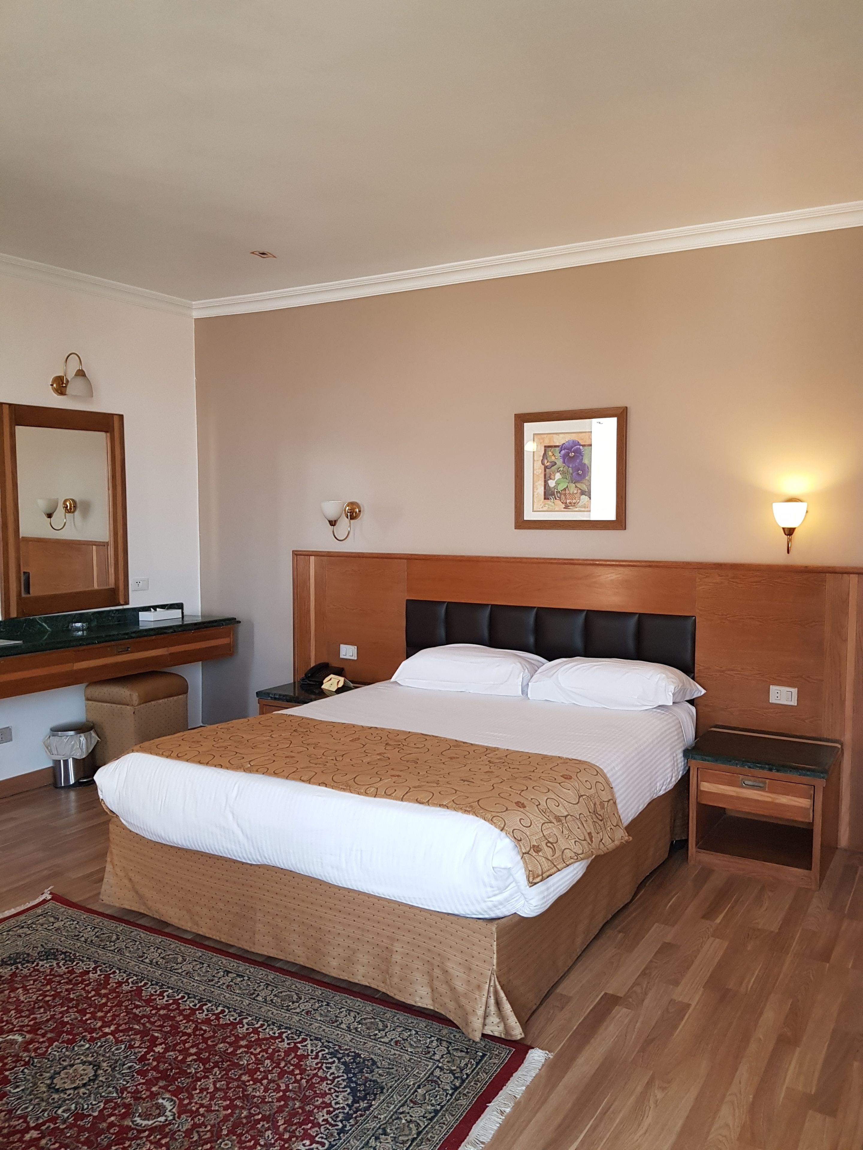 Habitación de hotel con cama, mesita de noche, tablero lateral, cabecero de madera, lámpara de pared e imagen enmarcada.
