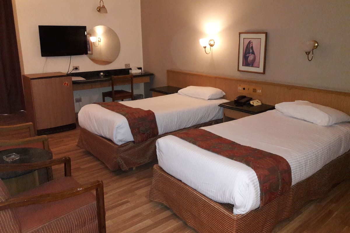Quarto de hotel com duas camas de solteiro, uma escrivaninha com cadeira, televisão e luminária na parede.