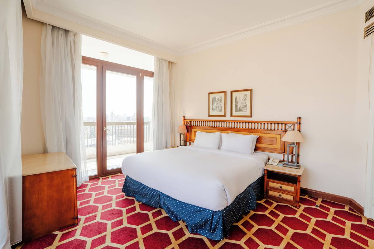 A hotel room with a large bed, a wooden nightstand, a desk, and a view of the city through the window.
