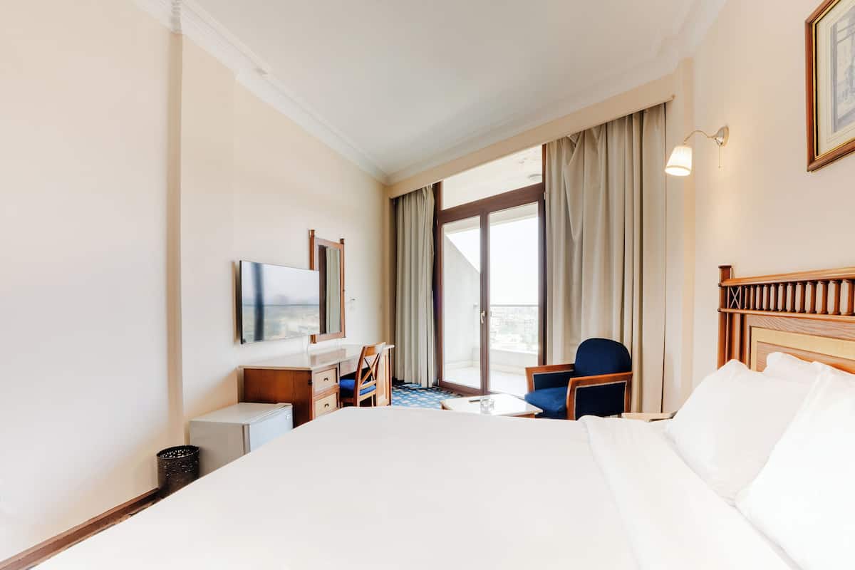 A hotel room with a large bed, a desk with a chair, a television, and a balcony with curtains.