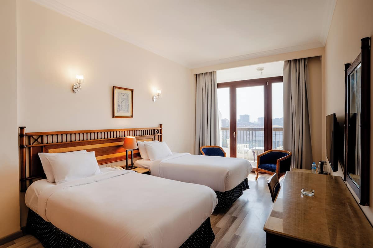 A hotel room with two beds, a wooden headboard, a dining table, a chair, a TV, and a city view.