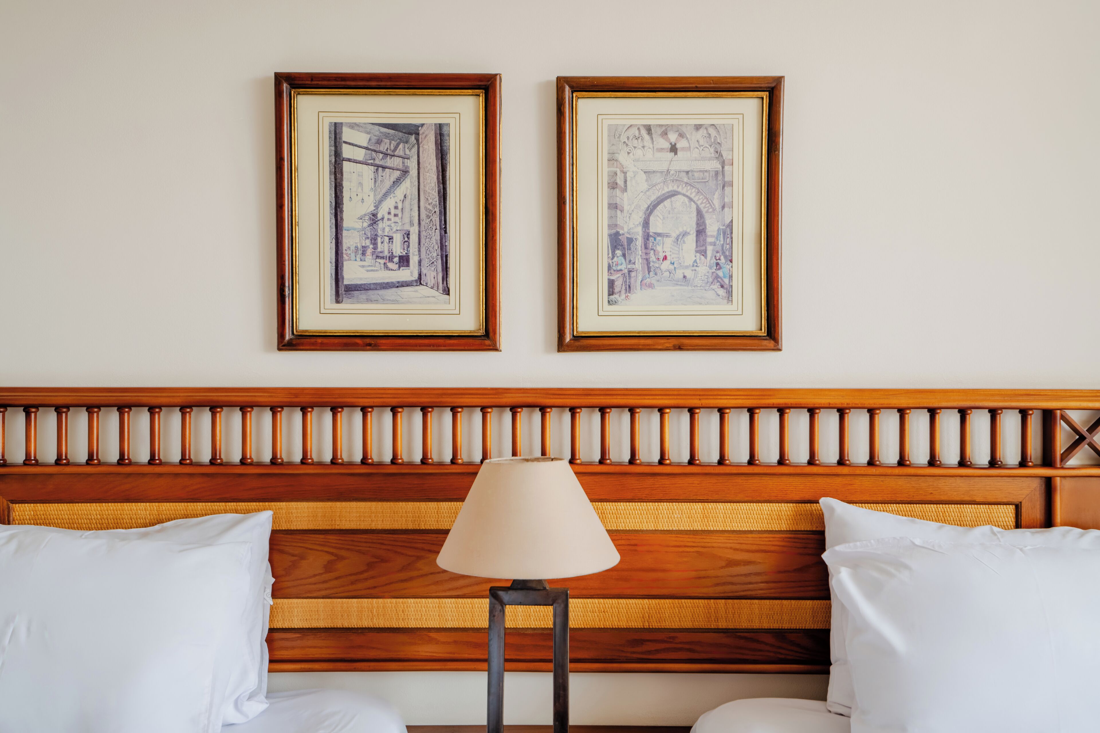 Habitación de hotel con cabecera de madera, dos cuadros en la pared, una lámpara de noche y dos almohadas blancas.