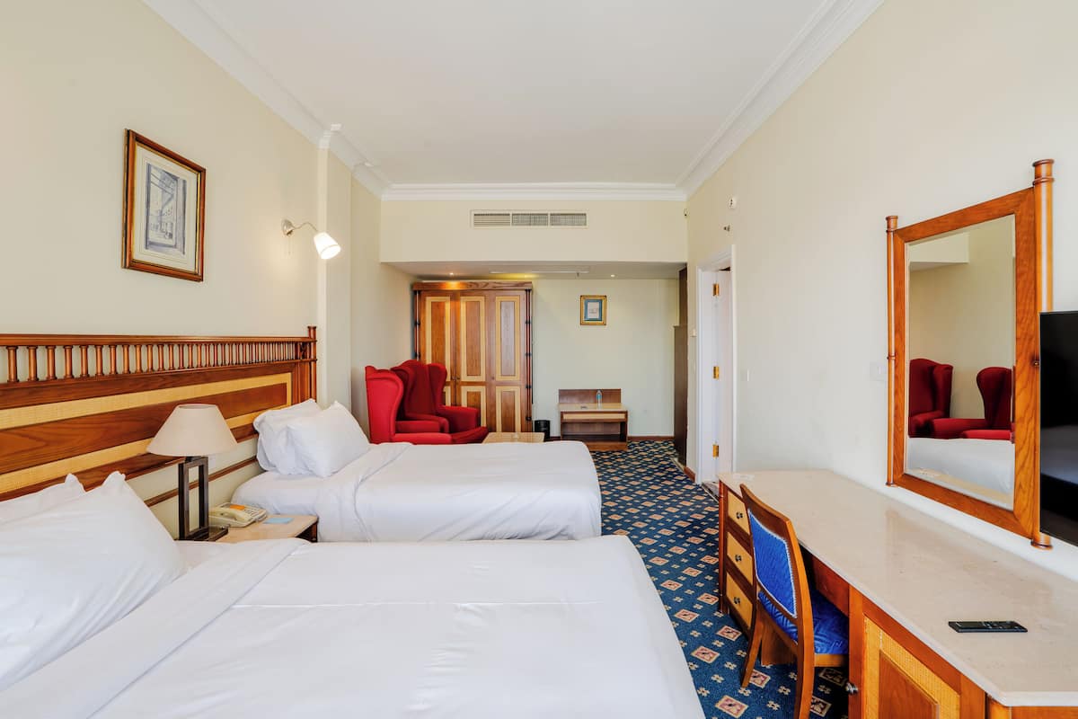 A hotel room with two beds, a desk, a chair, a mirror, a lamp, and a framed picture on the wall.