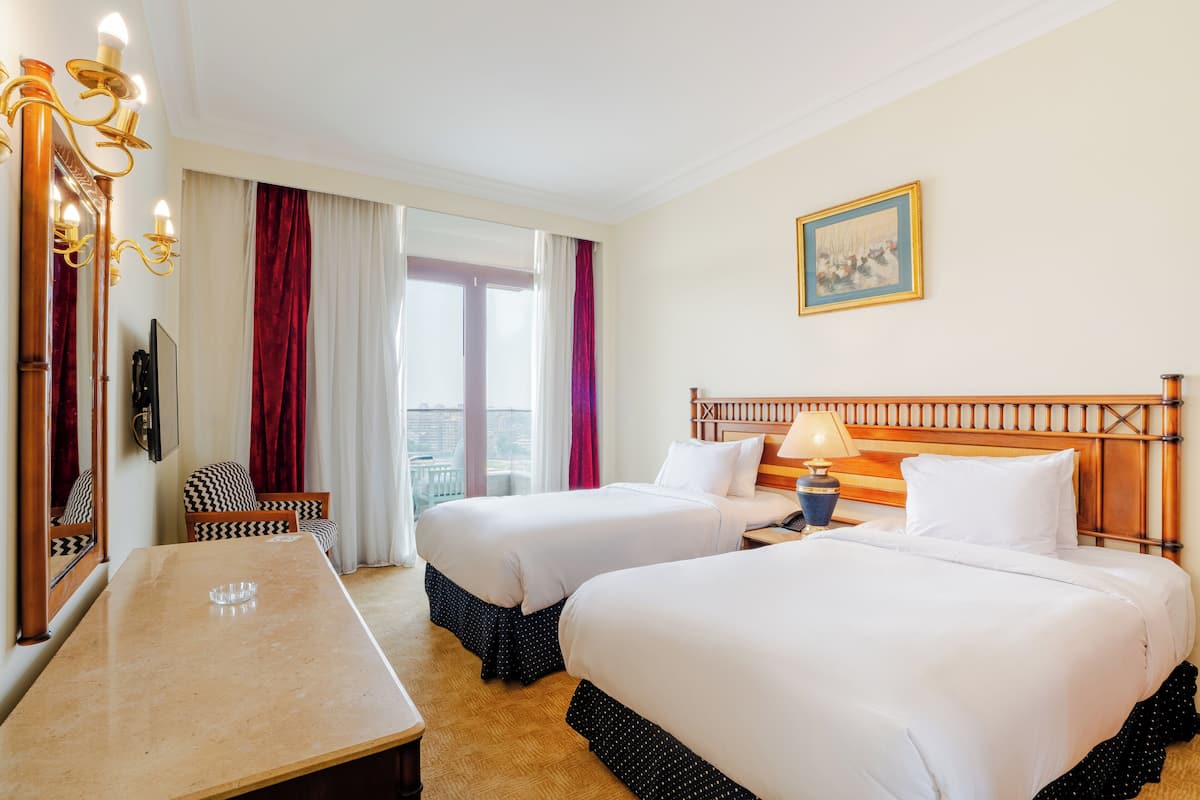 A hotel room with two beds, a desk, a chair, a lamp, and a painting on the wall.