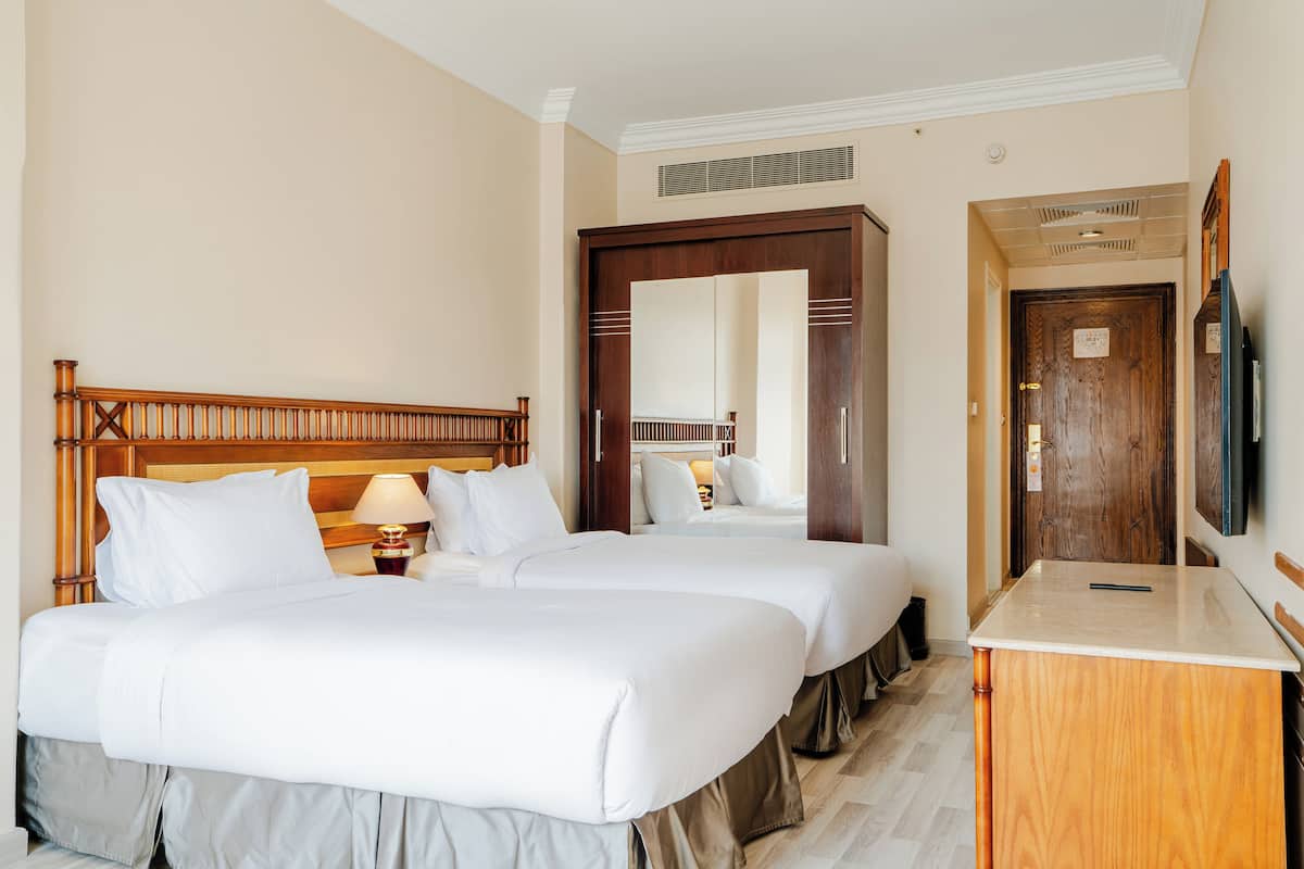 A hotel room with two beds, a wooden headboard, a bedside table with a lamp, a wooden dresser, and a television.
