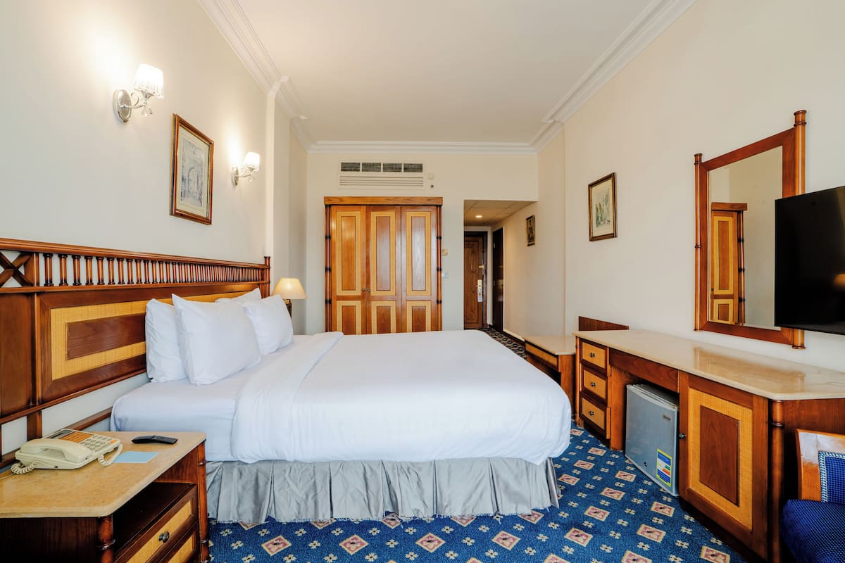 A hotel room with a large bed, wooden headboard, bedside table with a phone, a desk with a mirror, and a television.