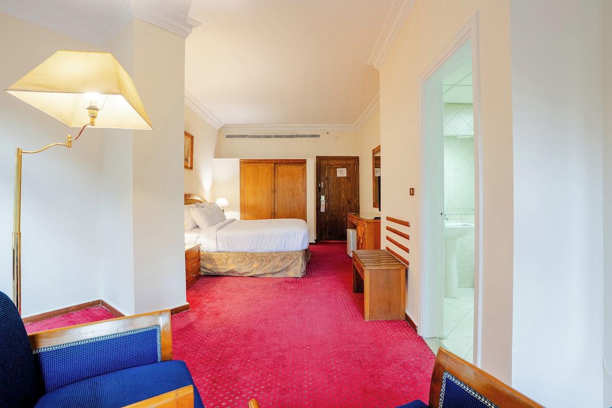 A hotel room with a bed, a desk, a chair, a lamp, and a door leading to a bathroom.