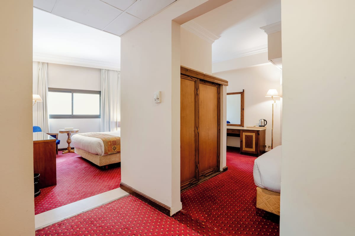 A hotel room with a red carpet, a bed, a desk with a lamp, a wardrobe, and a window with curtains.