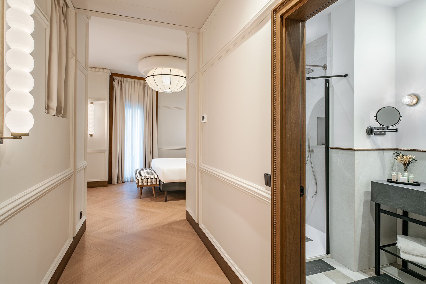 Pasillo de hotel con piso de madera, un banco, una cama y un baño con ducha e instalación de tocador.