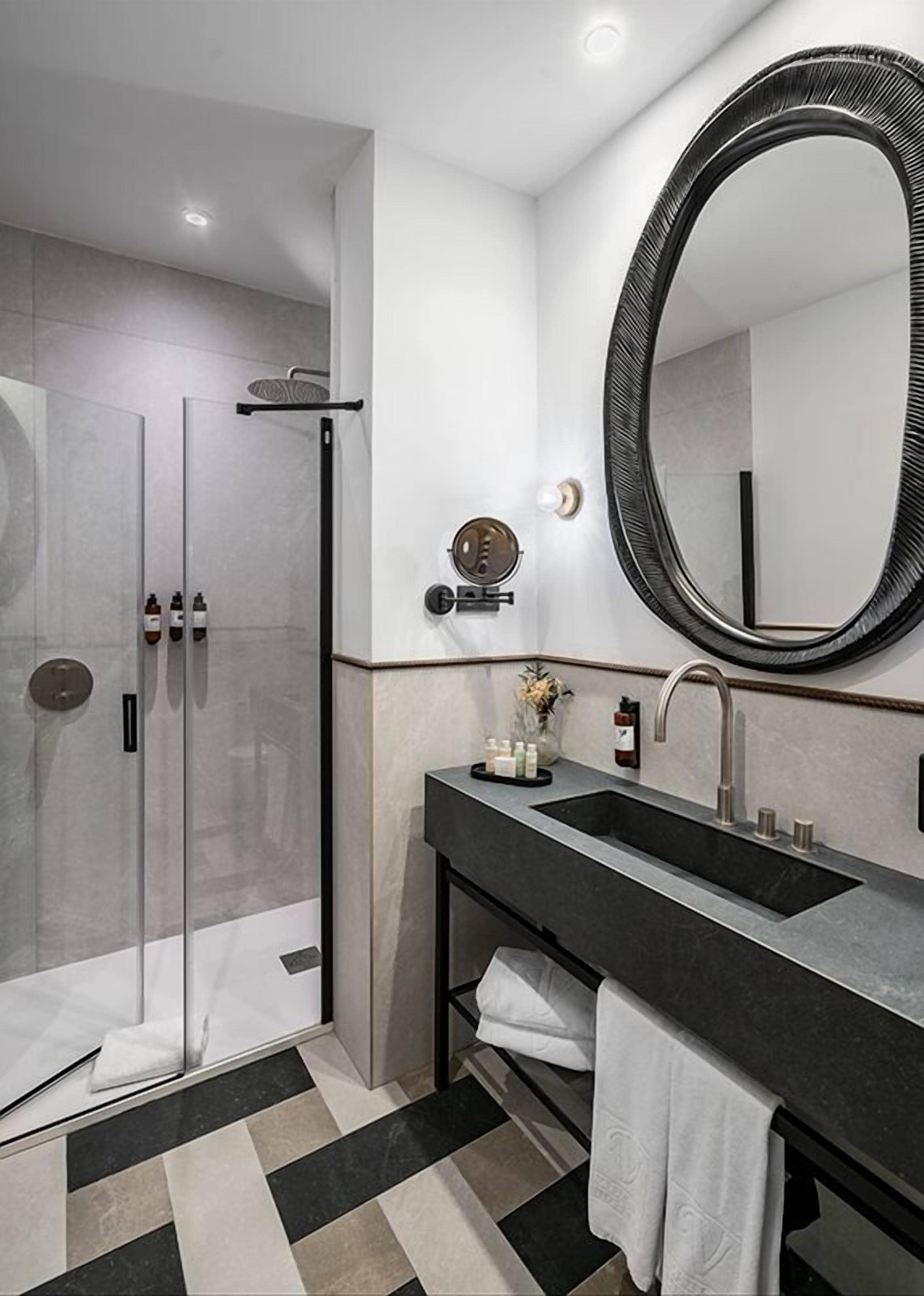 A modern bathroom with a glass-enclosed shower, a large oval mirror, and a black sink with a towel rack.